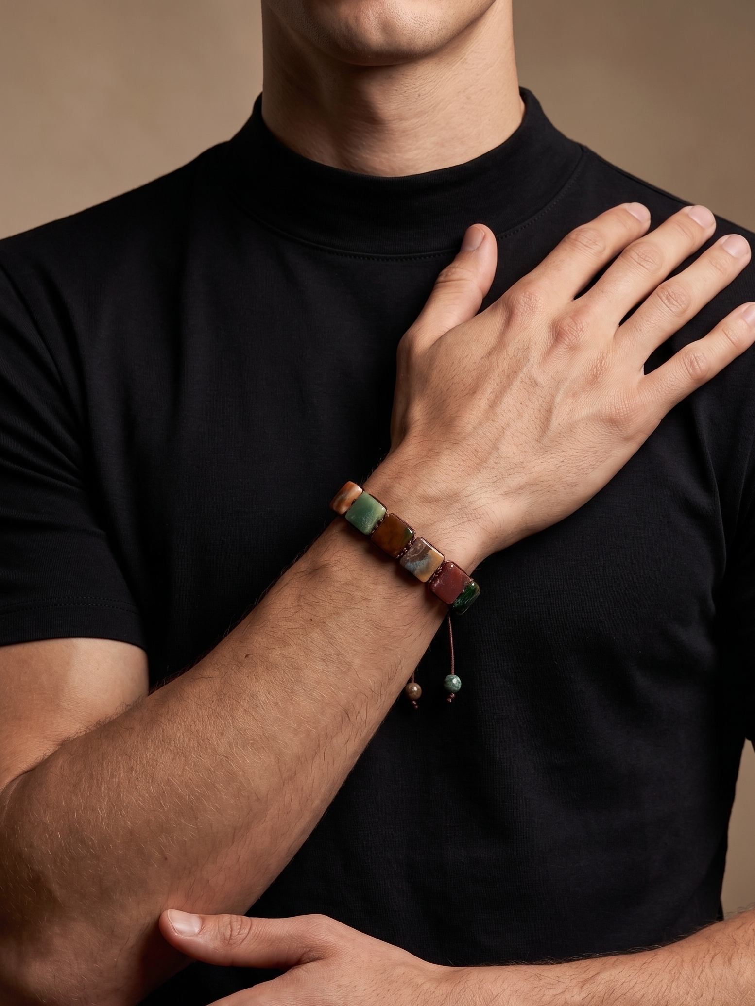 A close-up of a man wearing a black t-shirt and a multi-colored beaded bracelet on his wrist, posed with his hands crossed against a warm brown background.