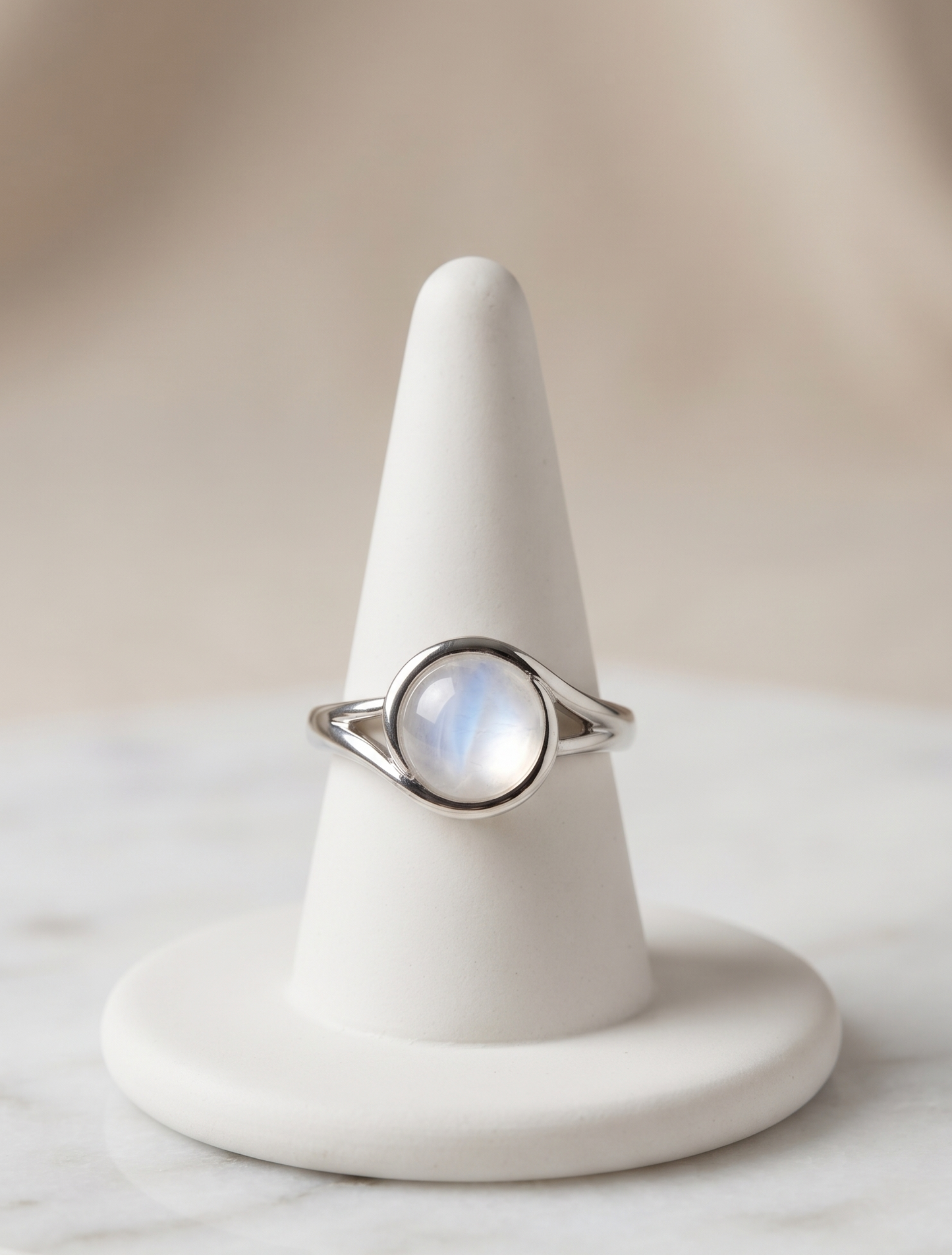 A silver ring with a round iridescent moonstone cabochon, displayed on a white cone jewelry stand over a light marble surface.