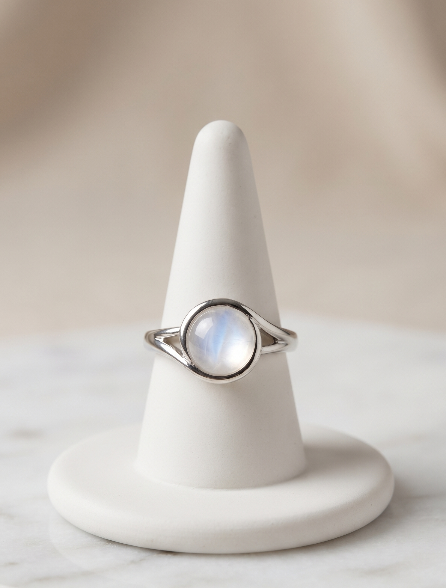 A silver ring with a round iridescent moonstone cabochon, displayed on a white cone jewelry stand over a light marble surface.