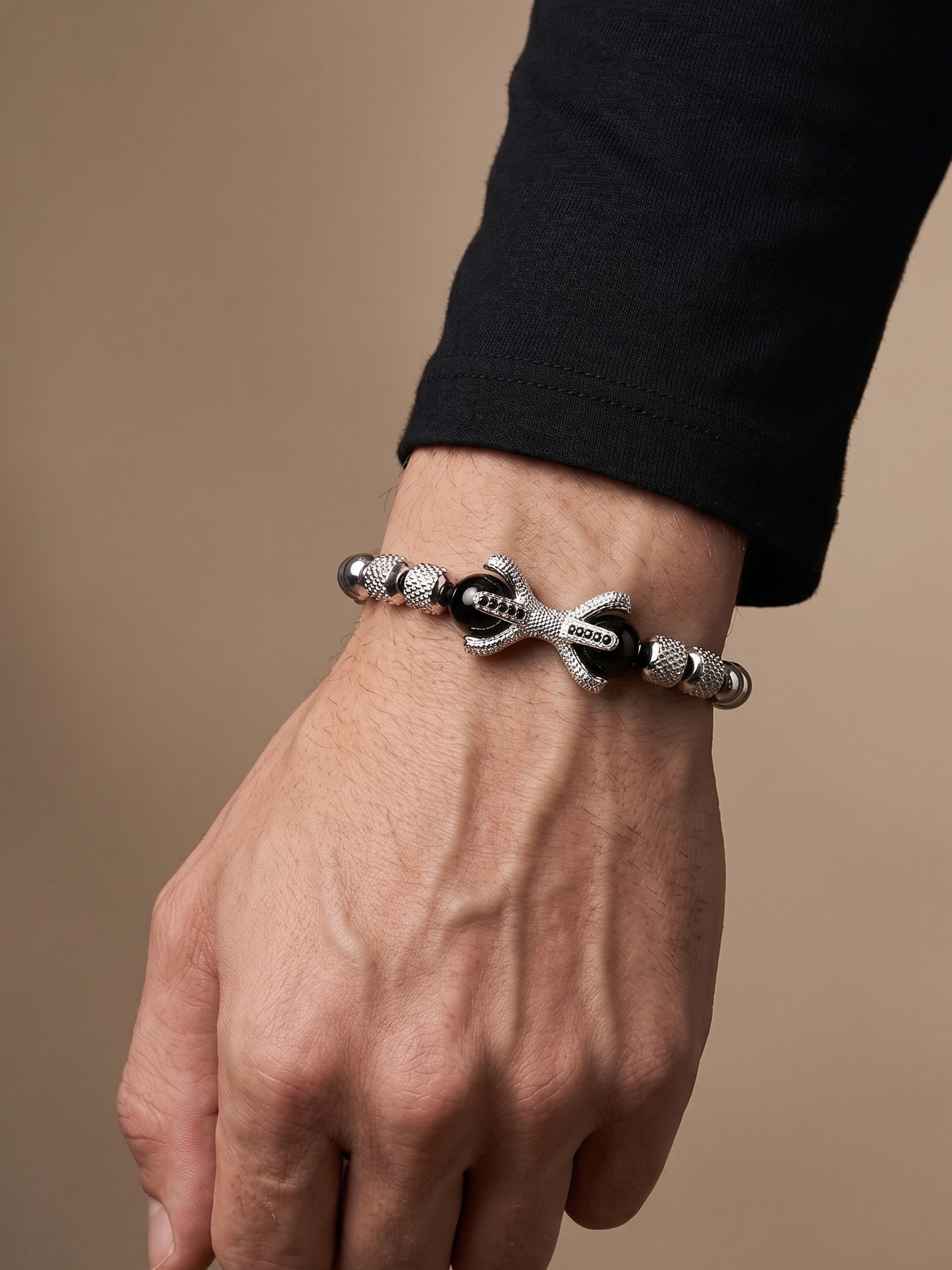A close-up of a person's hand and wrist wearing a silver-toned beaded bracelet with black accents and a central anchor charm. The person is wearing a black long-sleeve shirt, and the background is a solid warm beige studio backdrop.