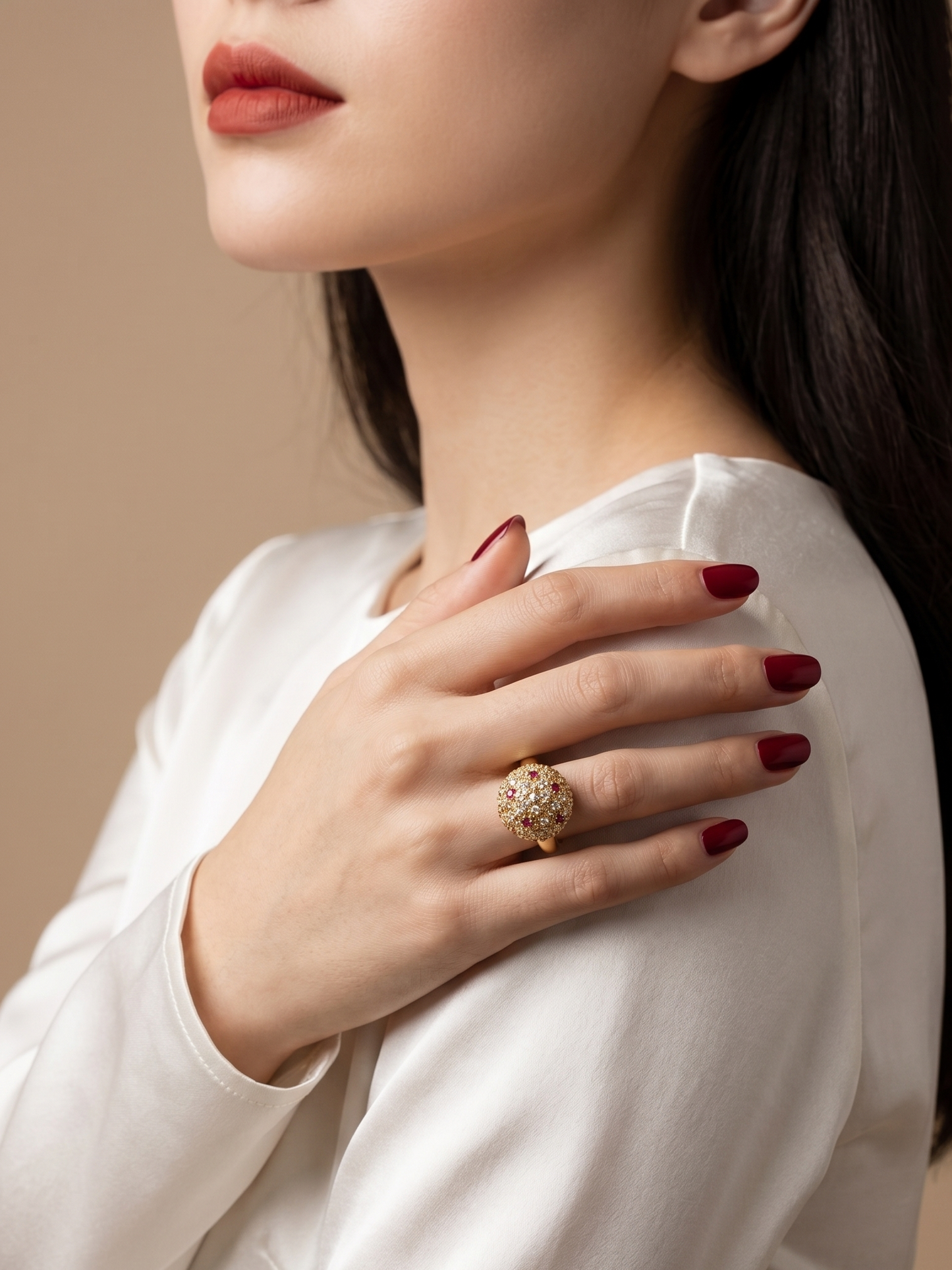 A close-up lifestyle shot of a woman wearing a large, ornate gold ring with a floral or leaf-like pavé design on her finger. She is wearing a white satin blouse and has dark red manicured nails, set against a solid warm beige background.