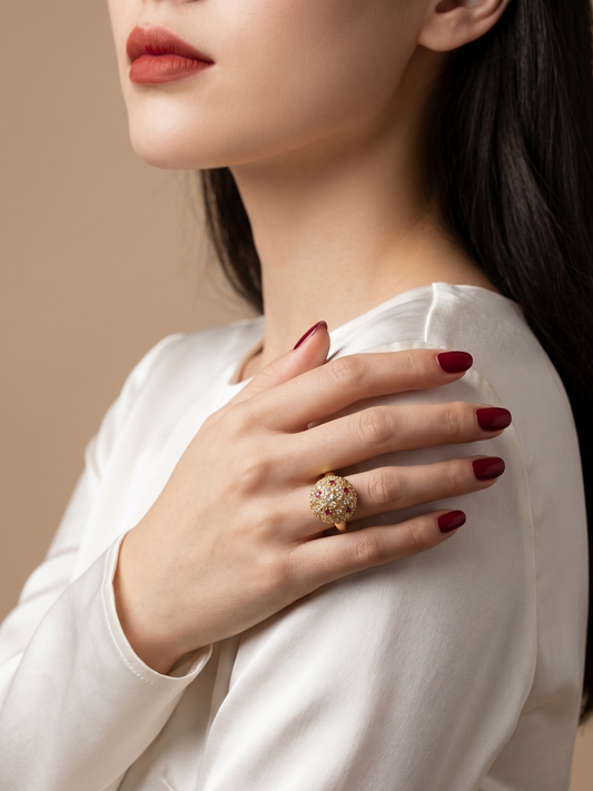 A close-up lifestyle shot of a woman wearing a large, ornate gold ring with a floral or leaf-like pavé design on her finger. She is wearing a white satin blouse and has dark red manicured nails, set against a solid warm beige background.