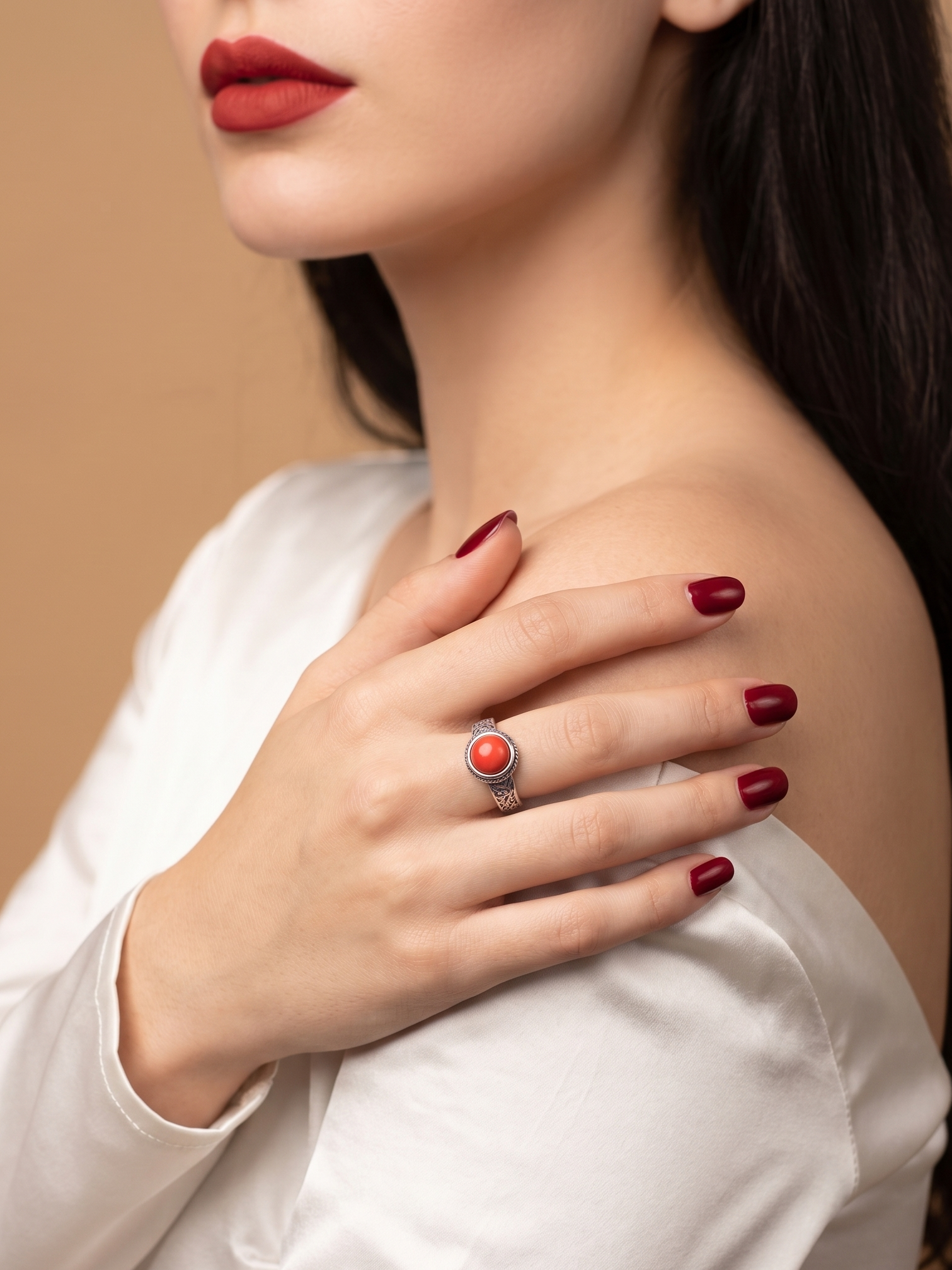 A close-up lifestyle shot of a woman wearing a silver ring with a round red cabochon stone, styled with dark red nail polish and a white satin top against a warm beige background.