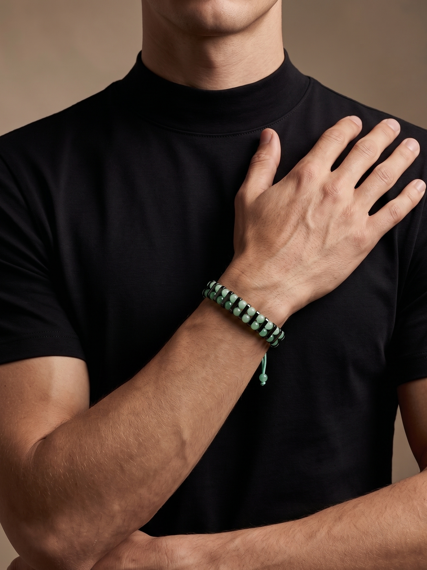 A close-up of a man wearing a black t-shirt and a green beaded bracelet on his wrist, with his hand resting on his chest against a warm beige background.