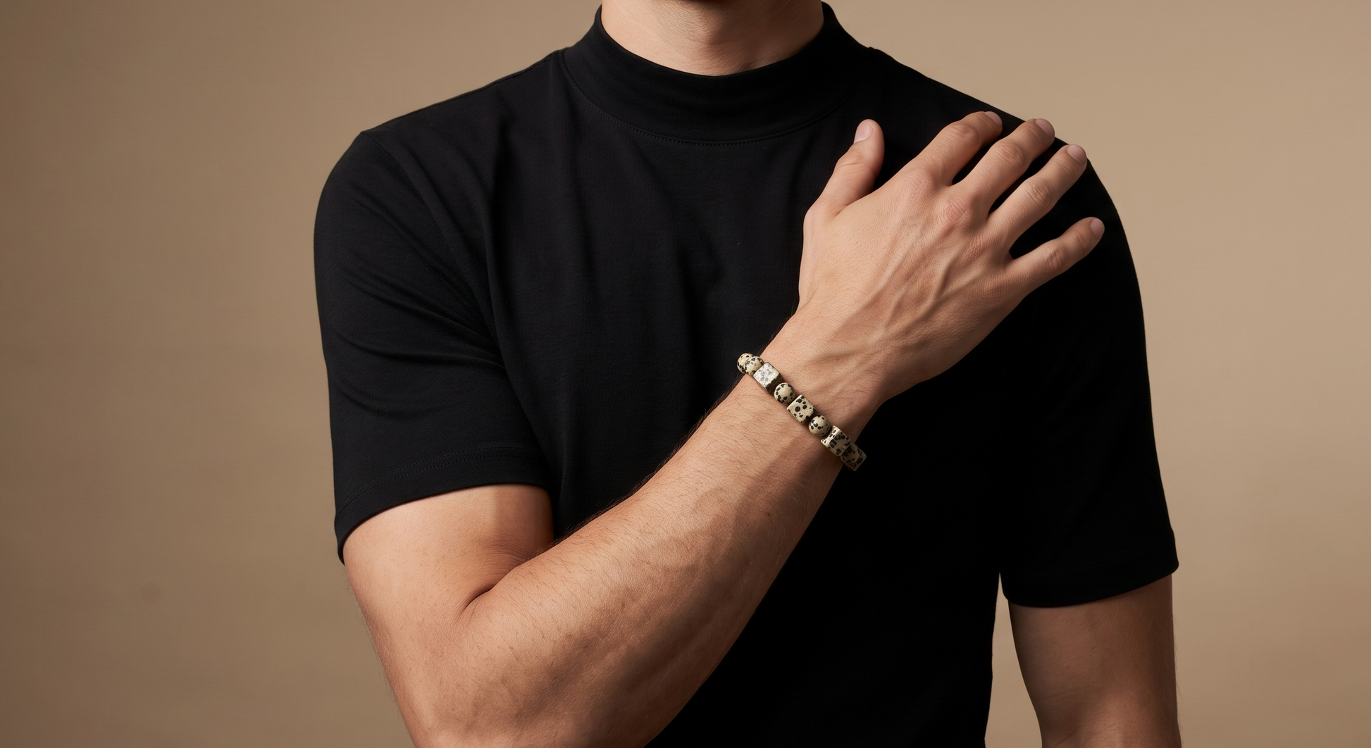 A man wearing a black short-sleeve mock neck t-shirt and a silver-toned beaded bracelet, posing with his hand on his shoulder against a solid warm brown background.