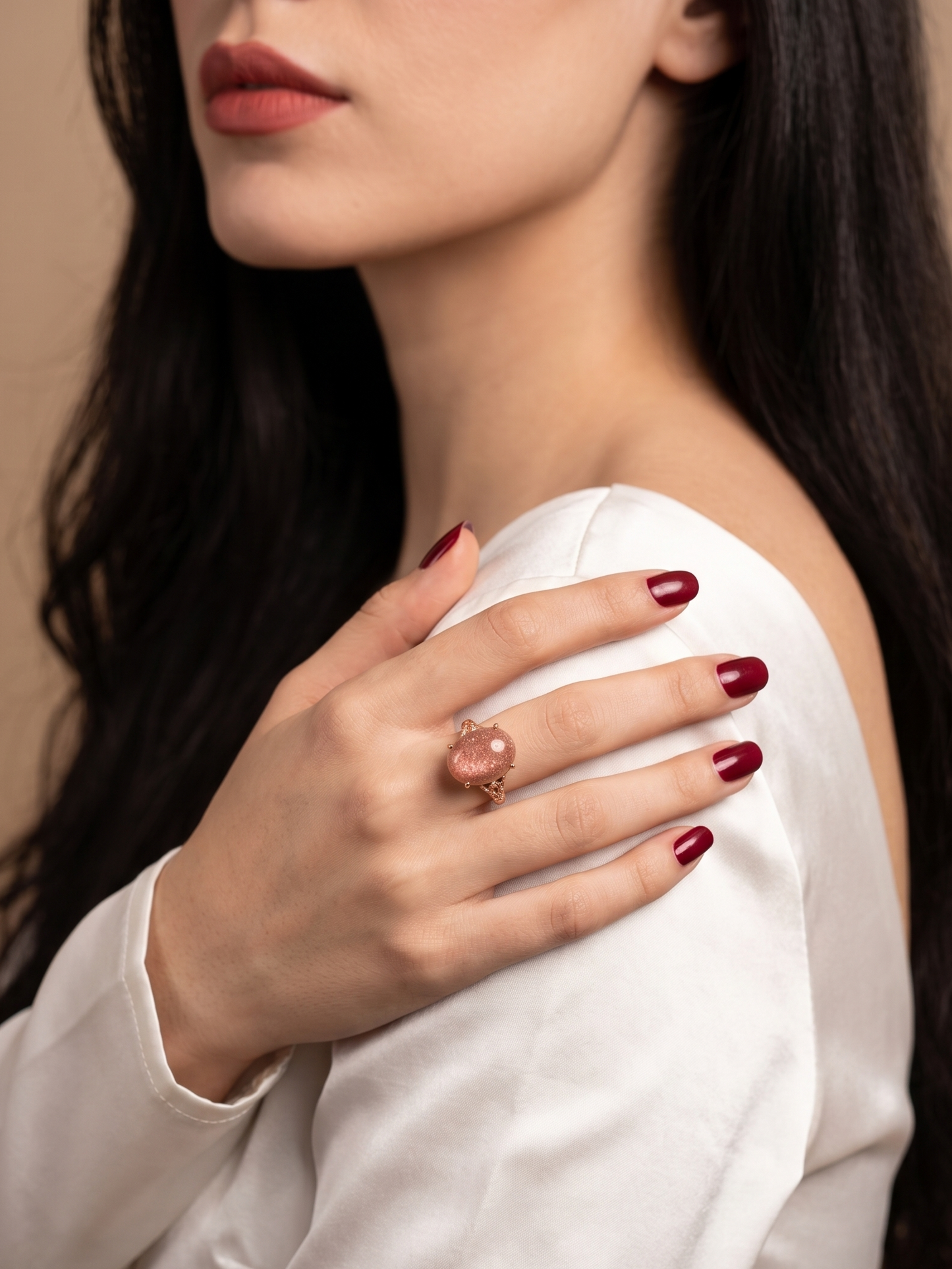 A close-up lifestyle shot of a woman wearing a pinkish-red oval gemstone ring on her finger, dressed in a white satin top against a warm beige background.