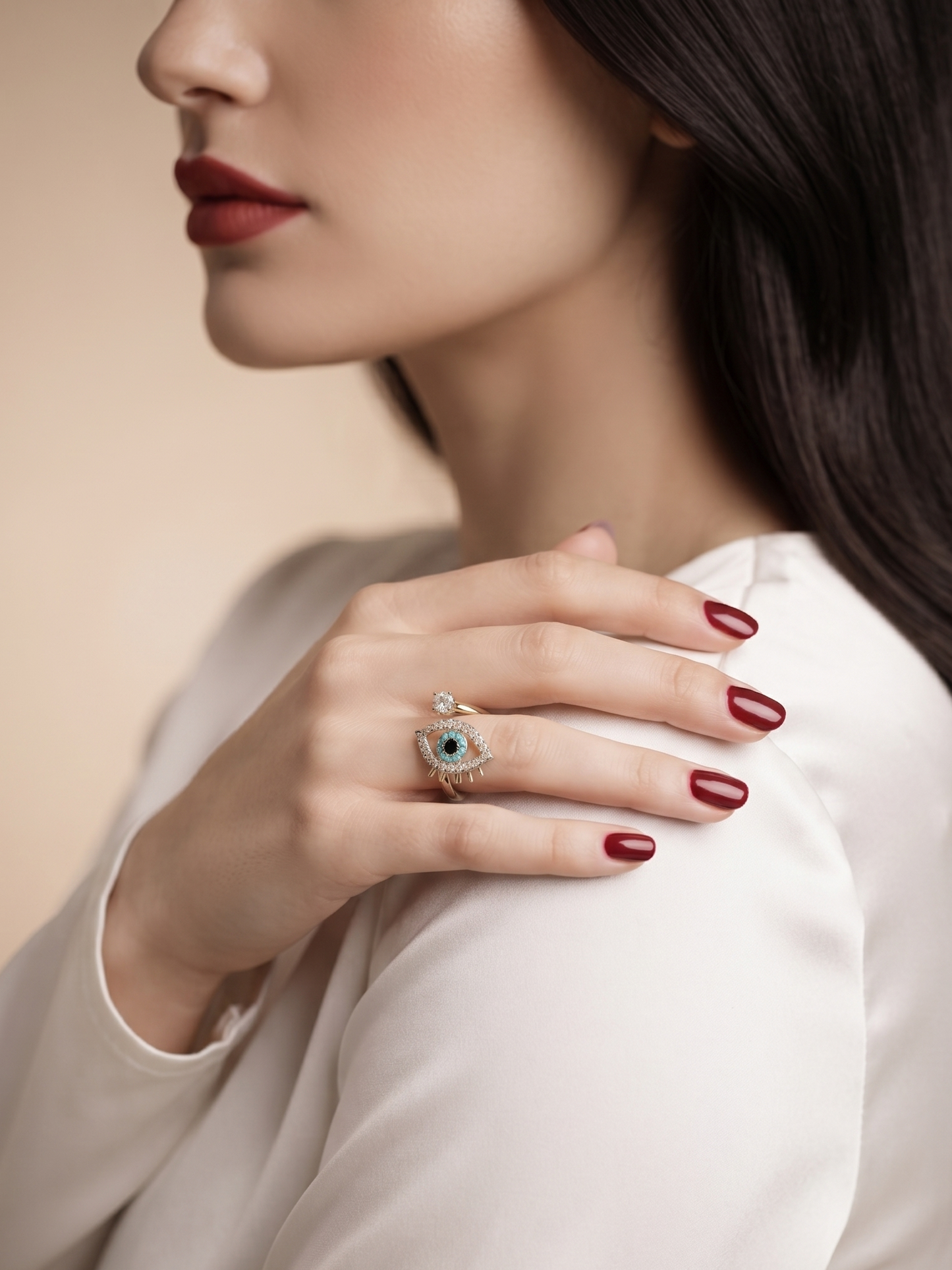 A close-up lifestyle shot of a woman wearing a silver-toned eye-shaped ring with a blue stone, styled with dark red manicured nails and a white satin blouse.