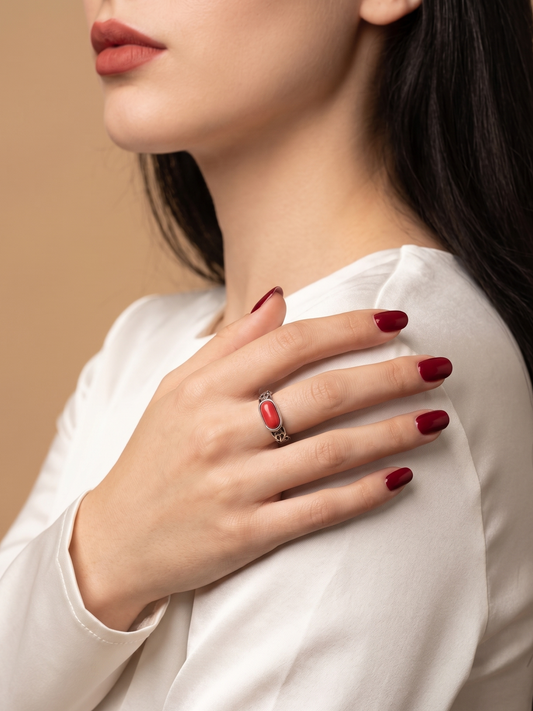 A close-up lifestyle shot of a woman wearing a silver ring with a red oval stone, resting her hand on her shoulder against a warm beige background.