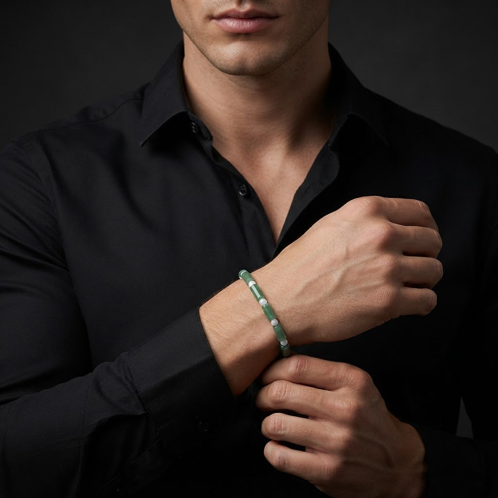 A man wearing a black button-down shirt adjusts his cuff while wearing a thin green beaded bracelet on his wrist, photographed against a dark studio background.