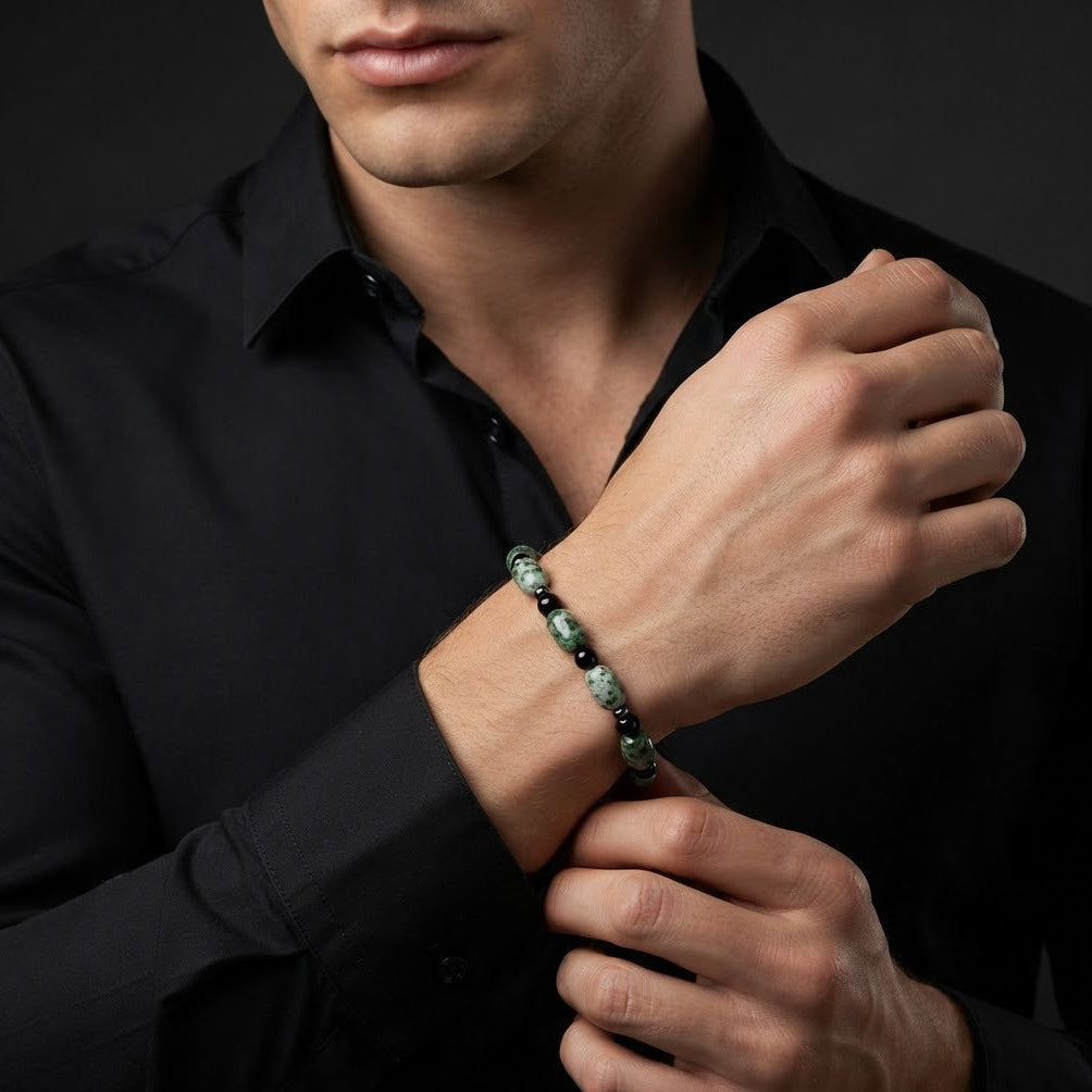 A close-up of a man wearing a dark green and black beaded bracelet on his wrist, dressed in a black button-down shirt against a dark studio background.