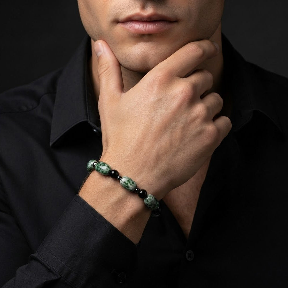 A close-up of a man wearing a beaded bracelet on his wrist, dressed in a black button-down shirt against a dark background.