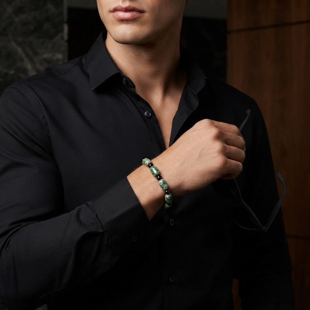 A close-up of a man wearing a black button-down shirt and adjusting his glasses while wearing a green beaded bracelet on his wrist.