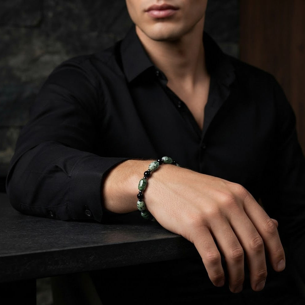 A man wearing a black button-down shirt rests his arm on a dark table, showcasing a beaded bracelet with green and black beads.
