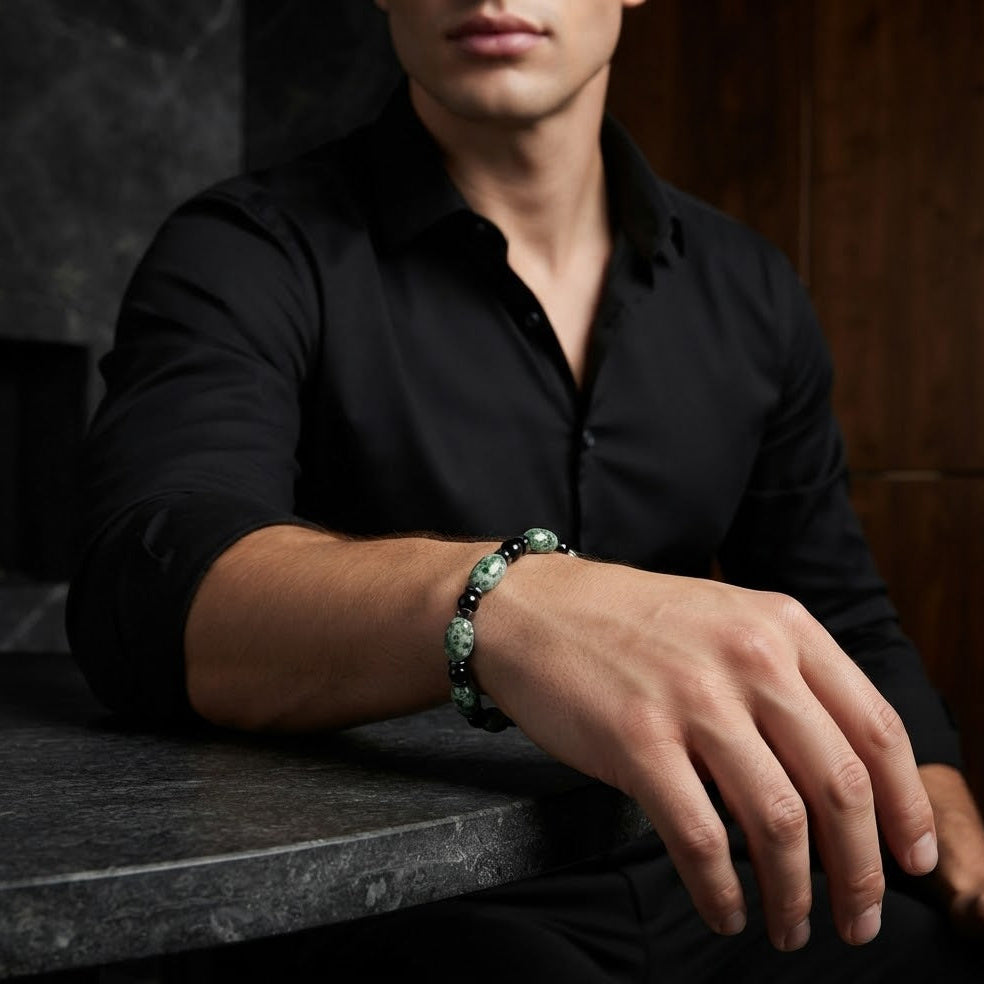 A man wearing a black button-up shirt and a beaded bracelet with green and black stones, resting his arm on a dark stone table.