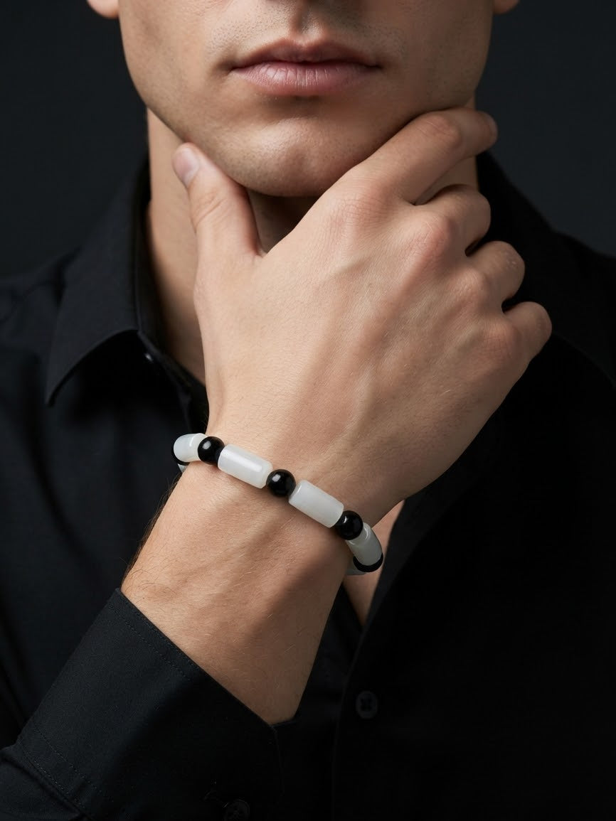 A close-up of a man wearing a beaded bracelet on his wrist. The bracelet features alternating white and black cylindrical beads, worn over a black collared shirt against a dark background.