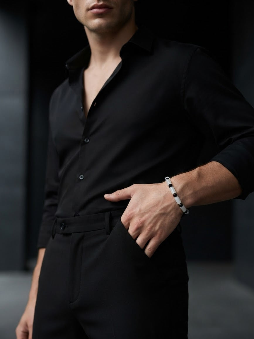 A man wearing a black long-sleeve button-down shirt and matching black trousers, with one hand in his pocket and the other wearing a white beaded bracelet, set against a dark, moody background.