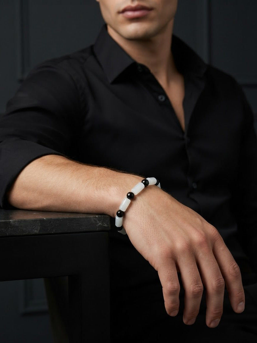 A man wearing a black button-down shirt and a white beaded bracelet with black beads, resting his arm on a dark table against a dark paneled wall.
