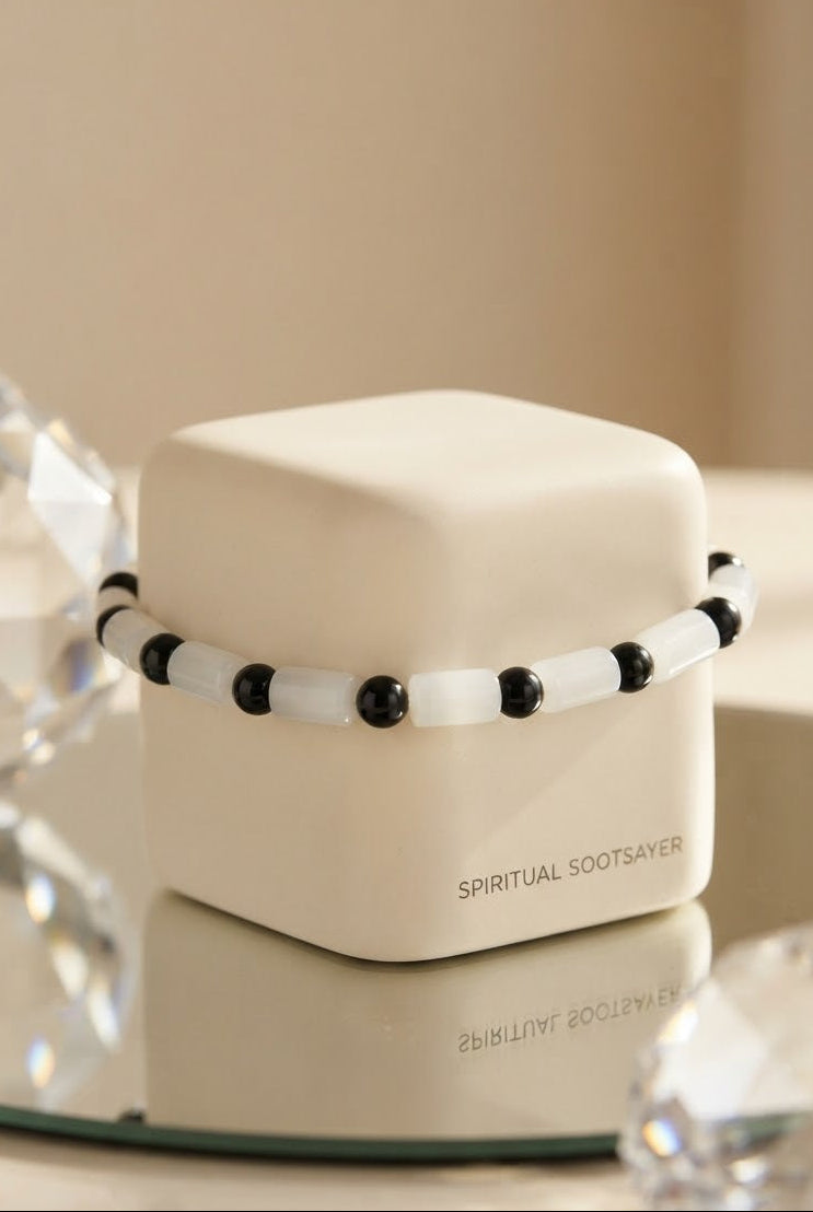 A beaded bracelet with alternating white and black beads displayed around a small cream-colored jewelry box. The box has the text 'SPIRITUAL SOOTSAVER' printed on the front.
