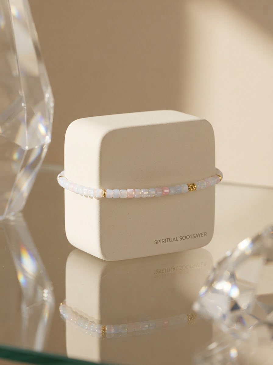 A delicate beaded bracelet with iridescent white and pale pink beads and small gold accents, wrapped around a matte cream jewelry box. The box has the brand name 'SPIRITUAL ROOTSAYER' printed on the front and is styled on a reflective glass surface with crystal props in the background.
