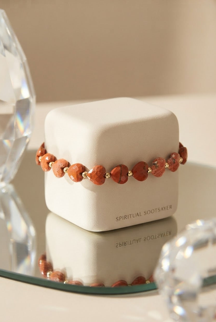 A beaded bracelet with terracotta-colored heart-shaped beads and small gold spacer beads, displayed on a white jewelry box. The box has the text 'SPIRITUAL SOOTSAVER' printed on it, and the scene is styled with clear faceted glass props on a reflective surface.
