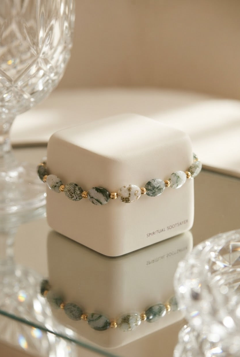 A beaded bracelet with green and white marbled beads and small gold spacer beads, displayed on a white jewelry box on a reflective glass surface.