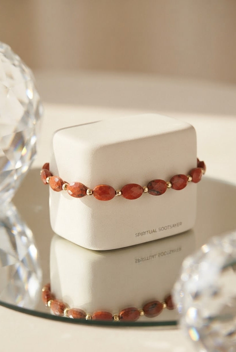 A beaded bracelet with reddish-orange oval beads and small gold spacer beads, displayed on a white jewelry box. The box is set on a reflective surface with blurred crystal spheres in the foreground.