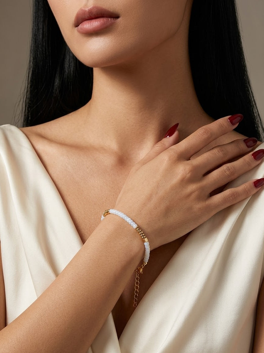 A close-up of a woman wearing a delicate white beaded bracelet with gold-toned chain details on her wrist, paired with a cream satin dress.