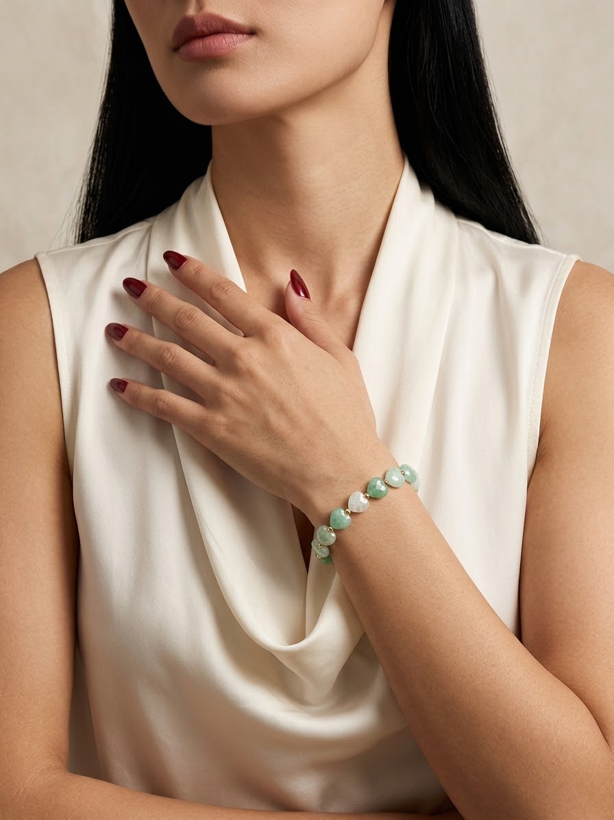 A close-up of a woman wearing a delicate beaded bracelet with light green and white stones, paired with a white satin top.