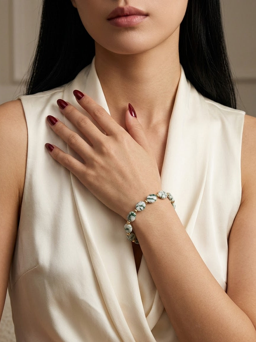 A close-up of a woman wearing a delicate beaded bracelet on her wrist, paired with a cream satin top. The bracelet features light blue and white beads with small gold-toned metal links.