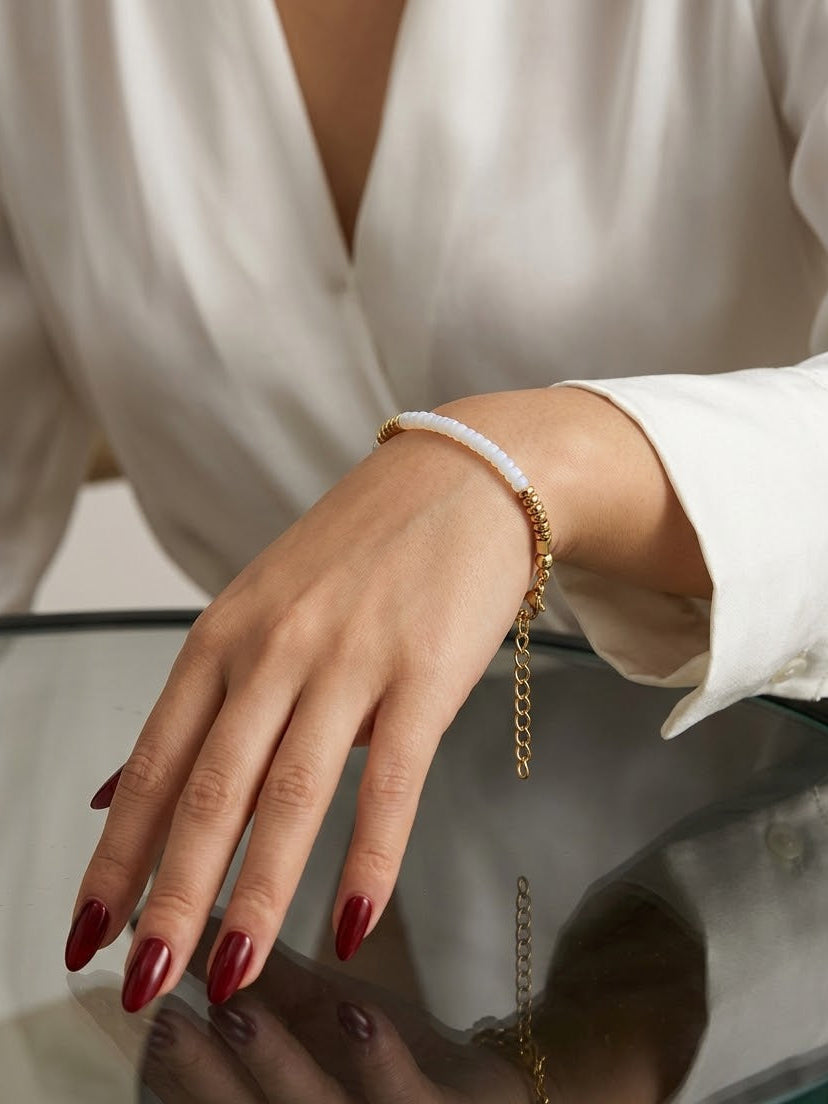 A close-up of a person's hand and wrist wearing a delicate gold chain bracelet with a white beaded section, styled with a white blouse and red manicured nails.