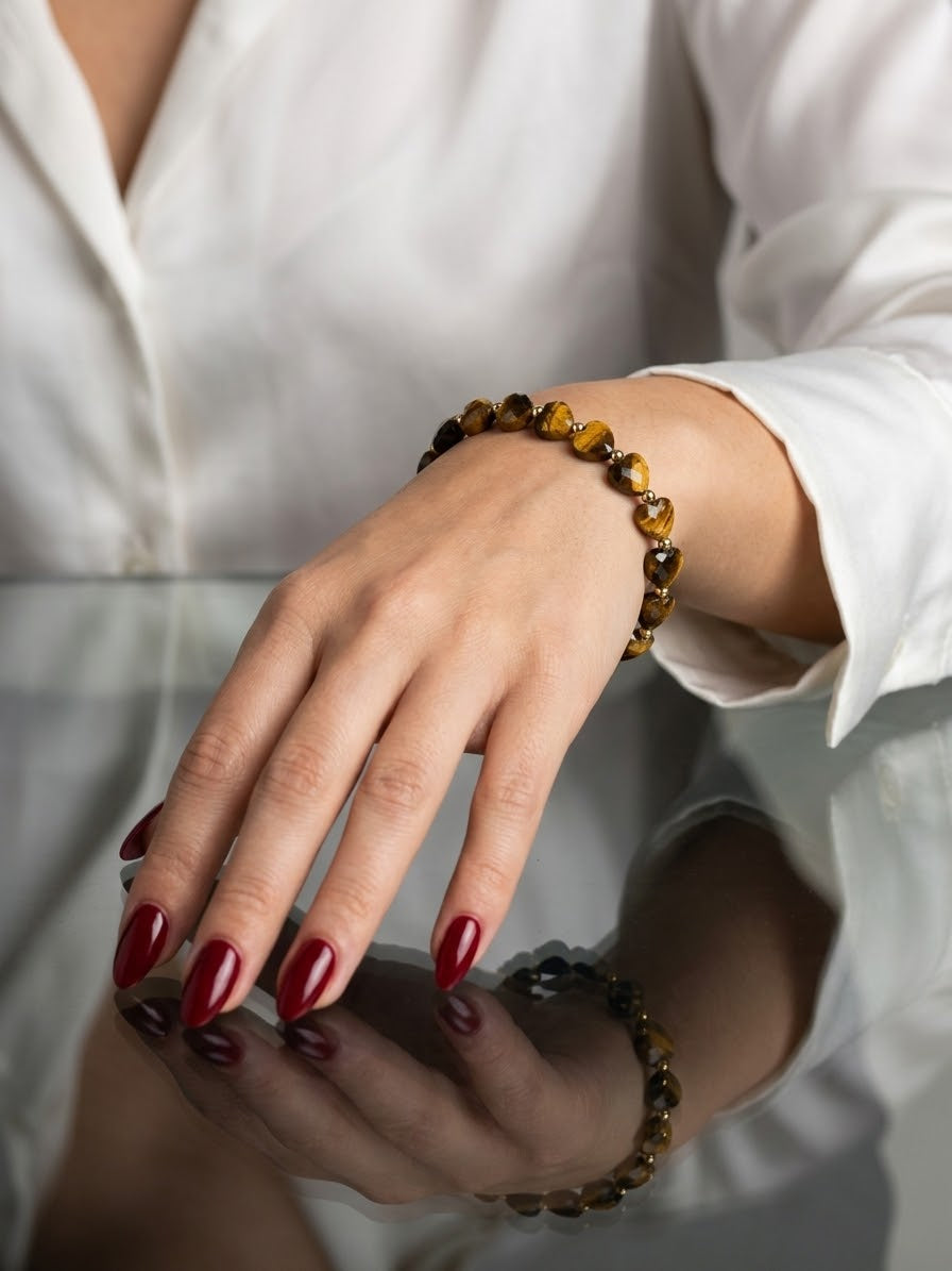 A close-up of a person's hand and wrist wearing a beaded bracelet made of polished brown and amber stones, with a matching bracelet reflected on a glossy surface. The person is wearing a white button-up shirt and has dark red manicured nails.