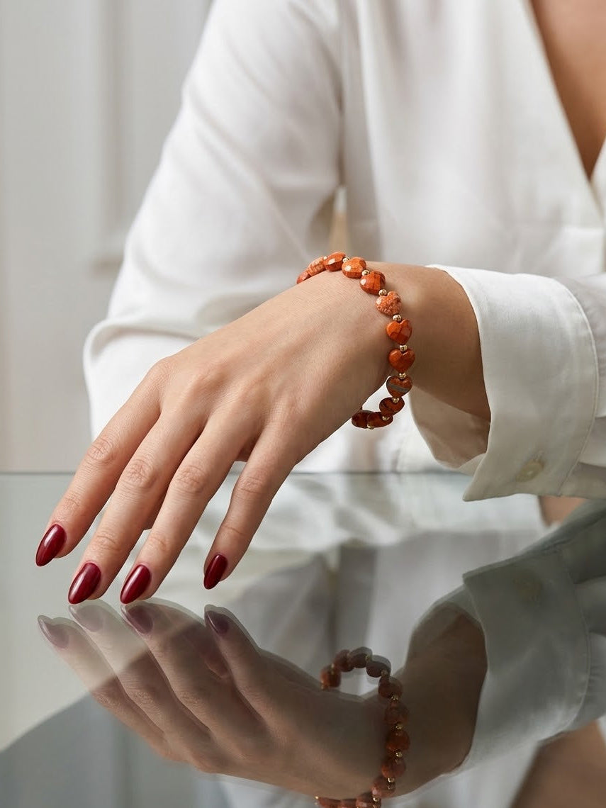 A close-up of a woman's hand wearing a beaded bracelet made of warm orange-brown polished stones, with her nails painted dark red. She is wearing a white long-sleeve blouse, and her hand is resting on a reflective glass surface.