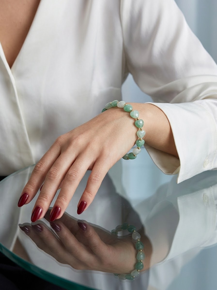 A close-up of a person's hand and wrist wearing a beaded bracelet made of light green and white polished stones, styled with a white blouse and red manicured nails.
