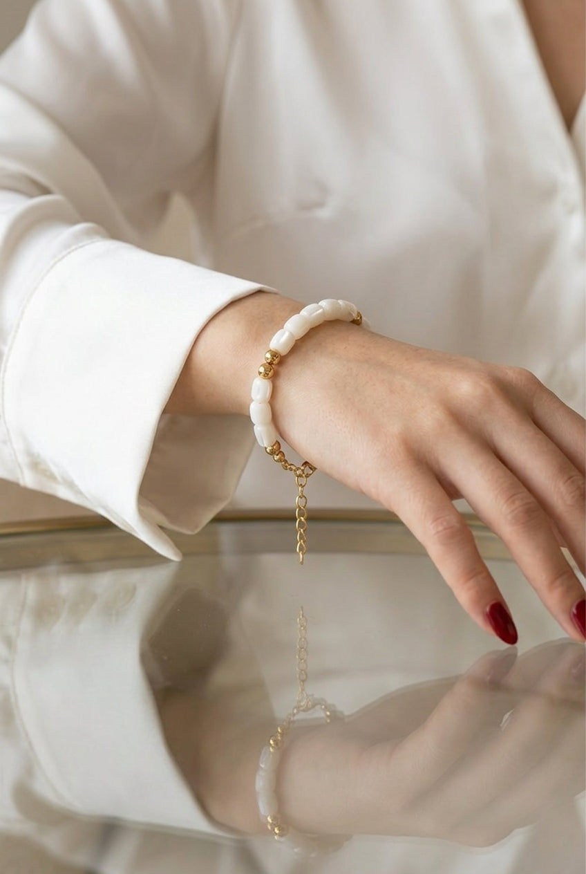 A close-up of a person's hand wearing a white beaded bracelet with gold spacer beads and a delicate gold chain charm, styled with a white long-sleeve blouse.