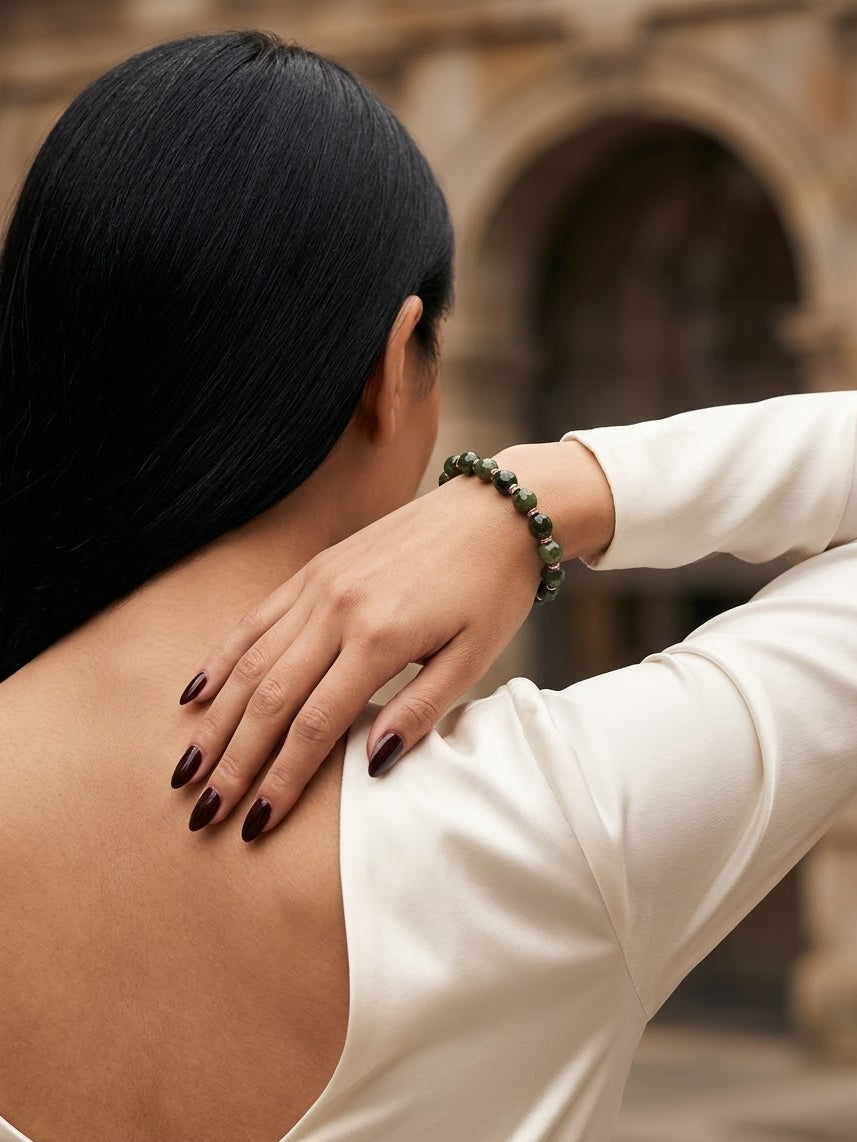 A woman with long black hair is shown from behind, wearing a dark green beaded bracelet on her wrist, with her hand resting on her shoulder. She is wearing a cream-colored long-sleeve top, and the background shows a blurred stone archway.