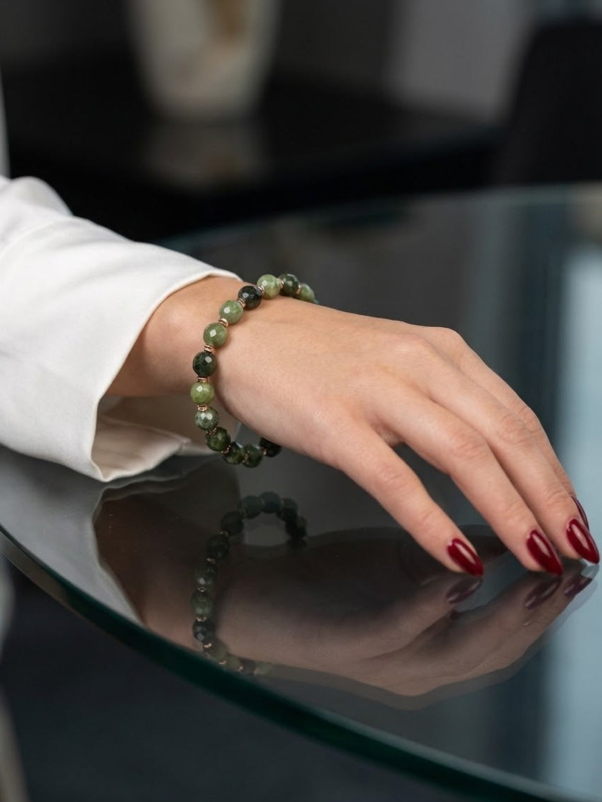 A close-up of a person's hand wearing a beaded bracelet made of dark green and olive stone beads, resting on a reflective dark glass table.