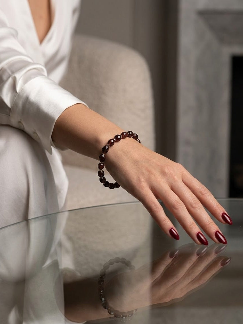A dark red beaded bracelet worn on a woman's wrist, shown in a lifestyle setting with her hand resting on a reflective glass table.
