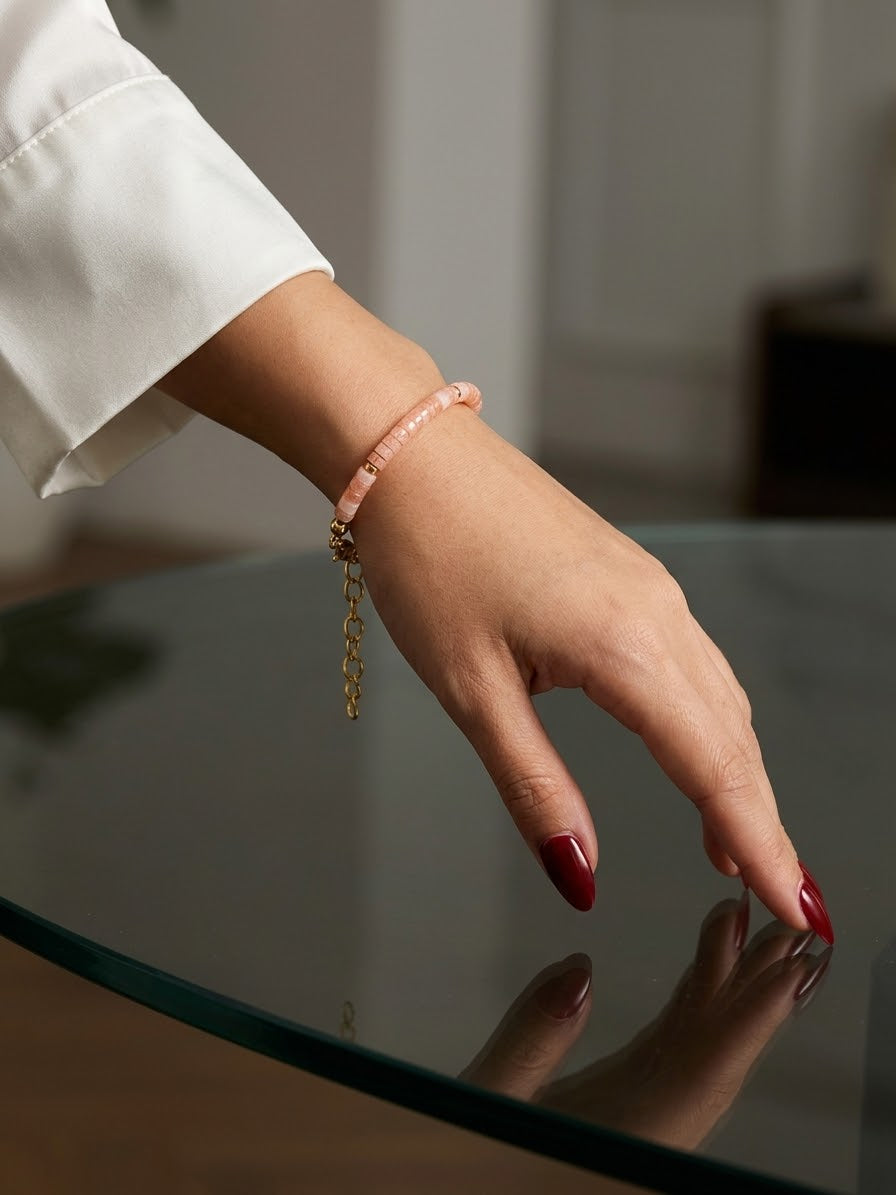 A close-up of a person's hand with dark red nail polish resting on a reflective glass table, wearing a delicate light pink beaded bracelet with a gold chain dangle.