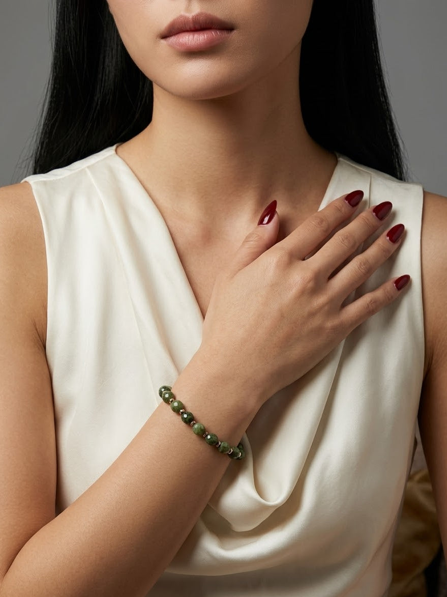 A close-up of a woman wearing a green beaded bracelet on her wrist, paired with a cream satin top. Her hand with dark red manicured nails rests on her chest against a neutral gray background.