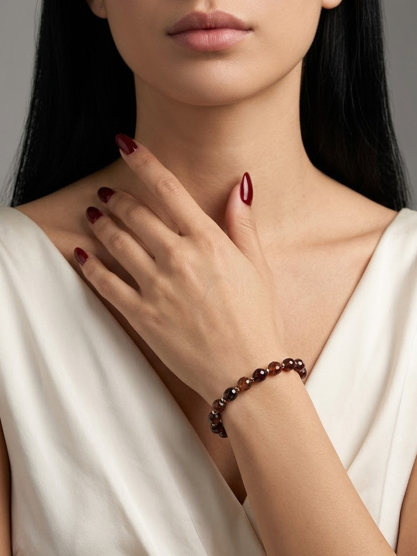 A close-up of a woman wearing a beaded bracelet on her wrist, with her hand resting near her neck. She is wearing a white satin top, and the background is a soft gray studio backdrop.