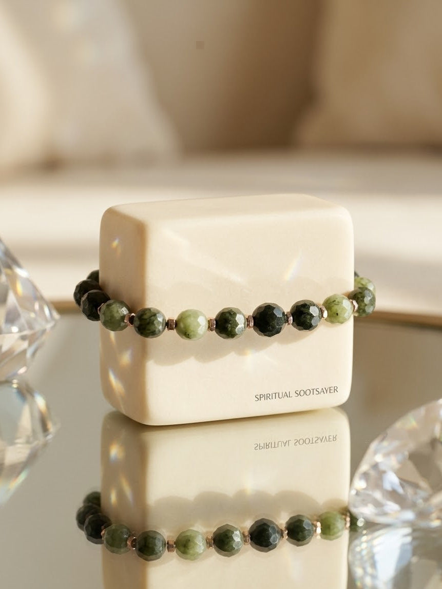 A beaded bracelet with dark green and light green faceted beads separated by small metallic spacer beads, displayed on a cream-colored cube. The brand name 'SPIRITUAL SOOTSAVER' is printed on the side of the cube.