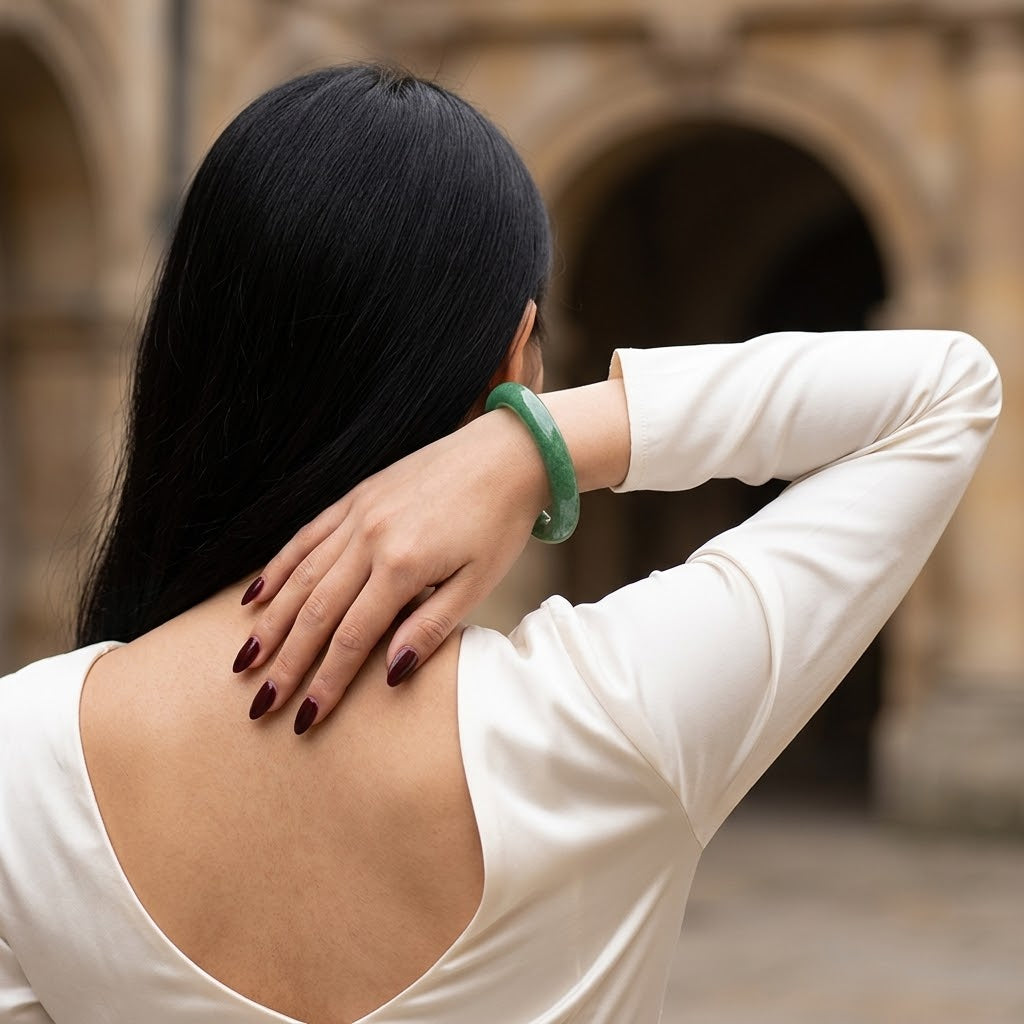 A woman with long black hair is shown from behind, wearing a white long-sleeve top and a green jade bangle on her wrist, with her hand resting on her neck. The background is a blurred stone building with arches.