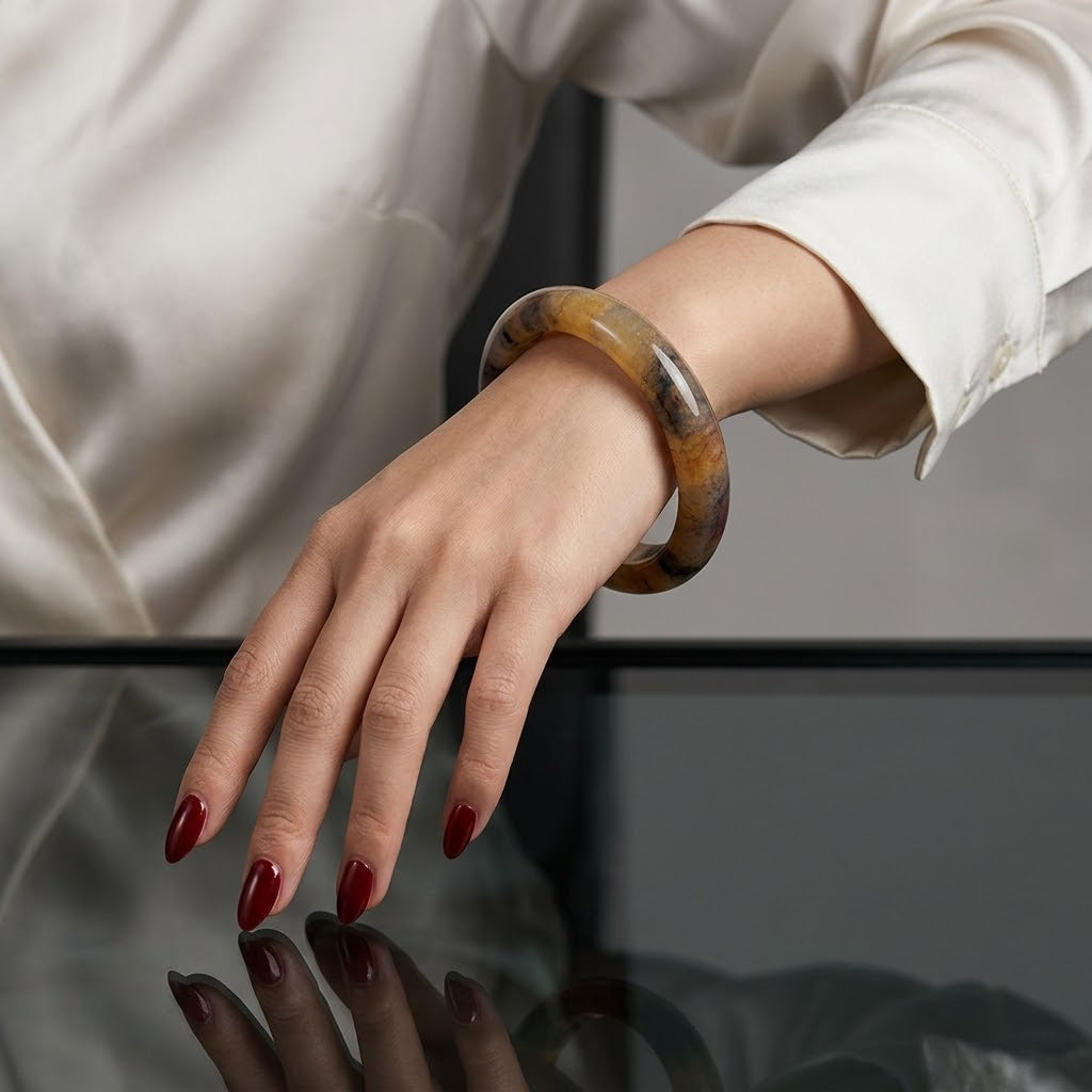 A close-up of a person's hand wearing a thick, polished tortoiseshell bangle bracelet, paired with a cream-colored satin blouse and dark red manicured nails.