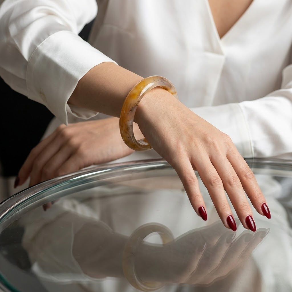 A person wearing a translucent amber-yellow bangle bracelet, shown from the chest down in a white blouse, resting their hand on a reflective glass table.