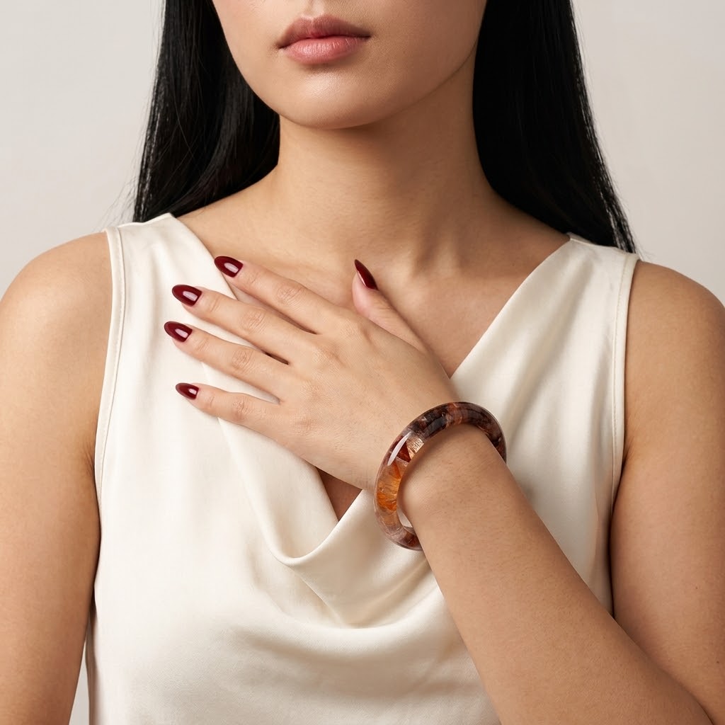 A close-up of a woman wearing a translucent tortoiseshell bangle bracelet on her wrist, paired with a cream satin top. The bracelet features a mottled pattern of dark brown, amber, and black.