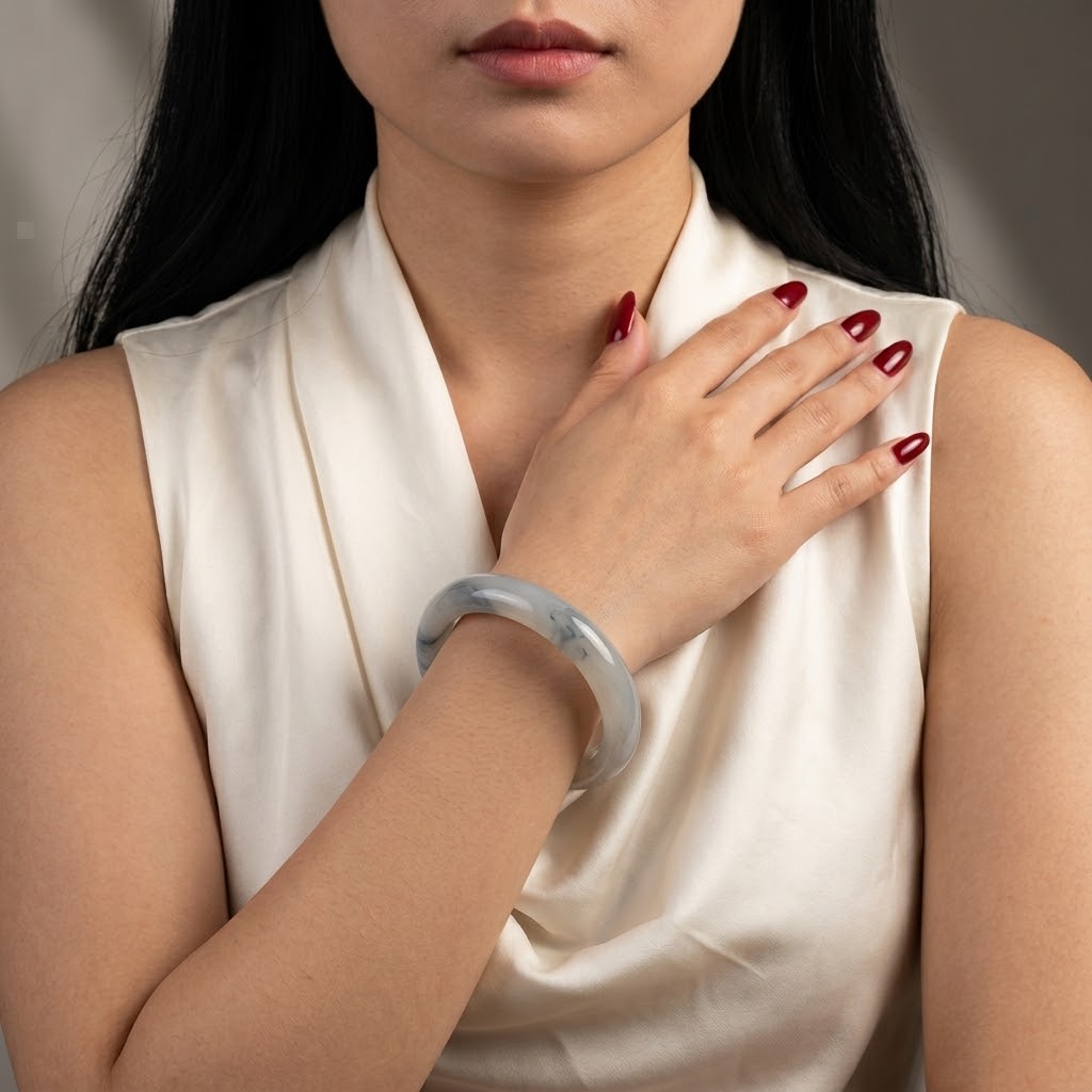 A close-up of a woman wearing a translucent pale gray-green jadeite bangle bracelet on her wrist, paired with a cream satin top. Her hand rests on her chest, showing dark red manicured nails.