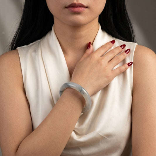 A close-up of a woman wearing a translucent pale gray-green jadeite bangle bracelet on her wrist, paired with a cream satin top. Her hand rests on her chest, showing dark red manicured nails.