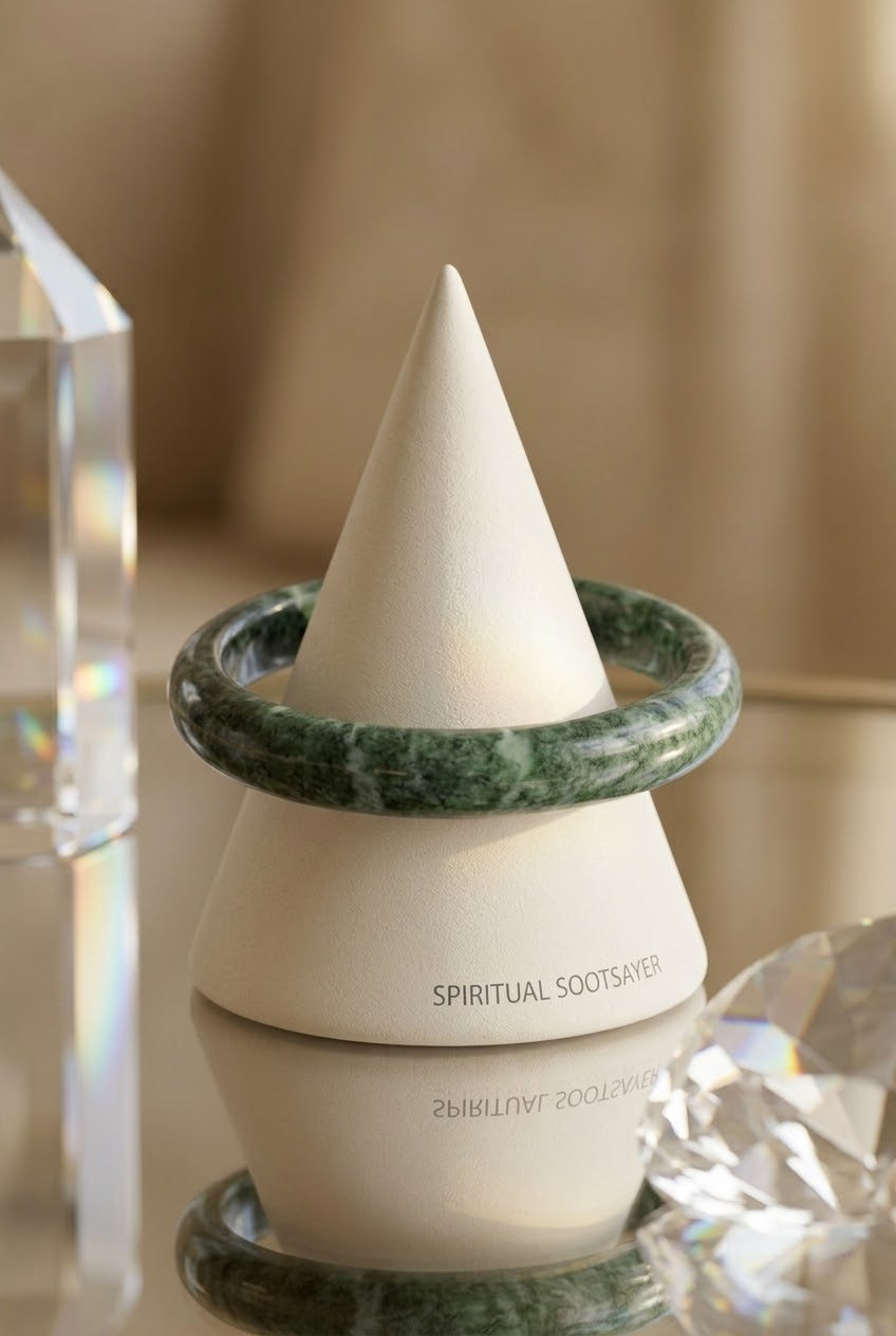 A white ceramic cone-shaped jewelry display stand with the text 'SPIRITUAL SOOTSAVER' printed on the front, surrounded by two green marble bangles, set against a soft beige background with decorative crystal props.