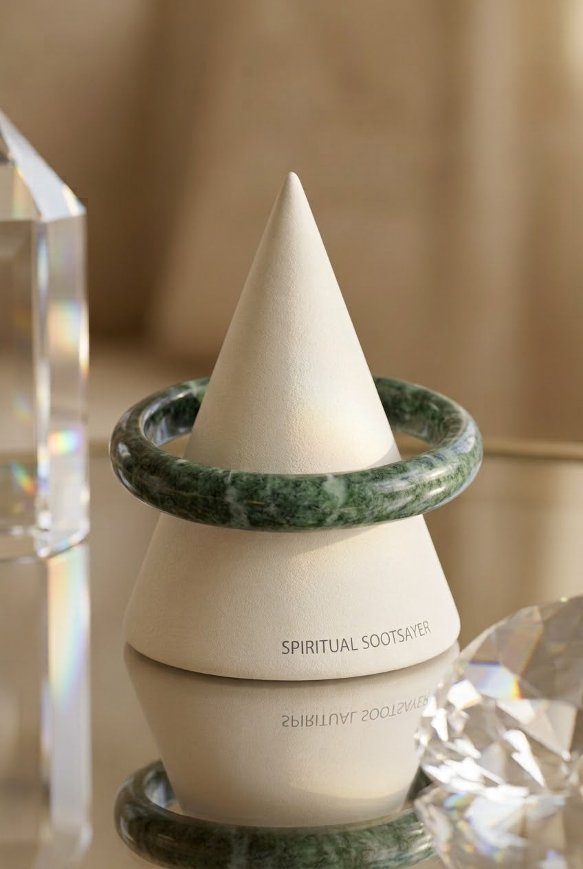 A ceramic cone-shaped holder labeled 'SPIRITUAL SOOTSAVER' in black text, with two green marbled bangles resting around its middle, set against a warm beige background with decorative crystal props.