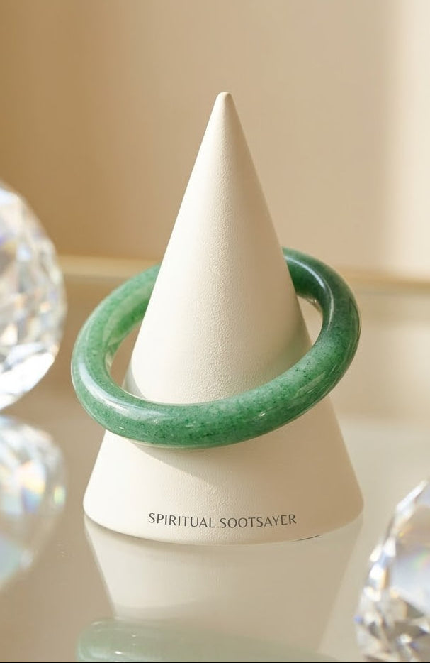 A green stone bangle bracelet displayed on a white cone jewelry stand, with the brand name 'SPIRITUAL SOOTSAVER' printed on the base. The scene is styled with clear faceted crystal spheres on a reflective surface against a warm beige background.